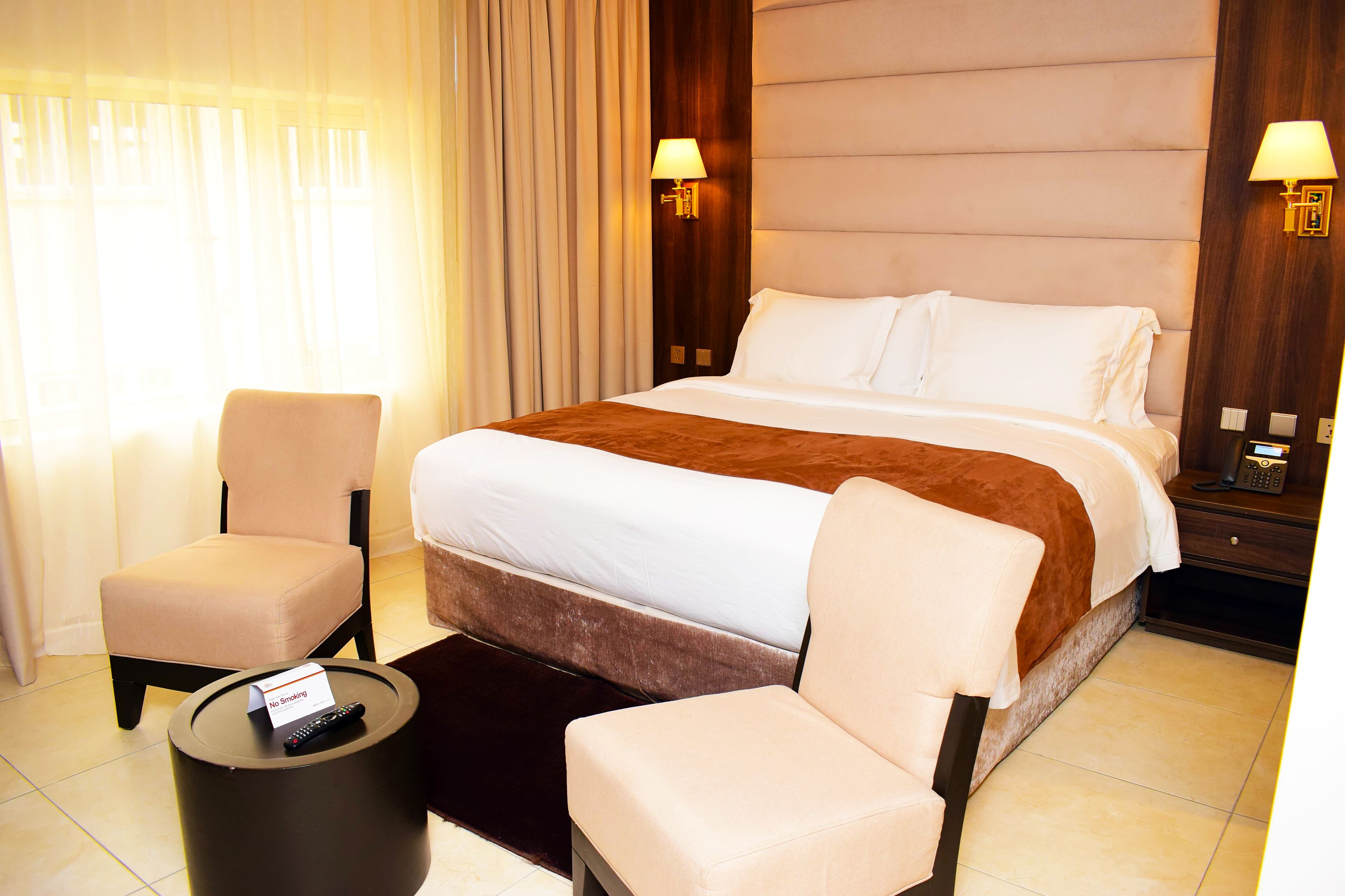 Une chambre d’hôtel avec un grand lit, deux fauteuils, une petite table ronde avec une télécommande et une lampe fixée au mur.