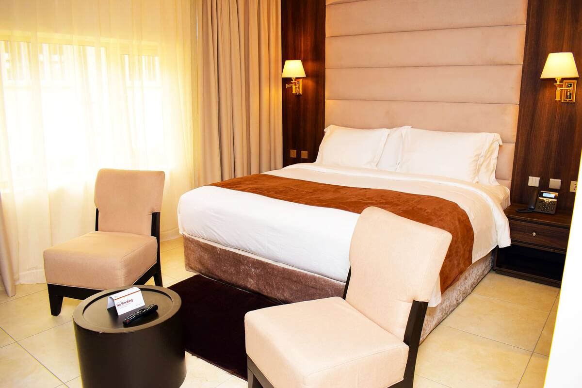 Quarto de hotel com uma cama grande, duas poltronas, uma mesa redonda pequena com um controle remoto e um aviso de 'Não Fumar'.