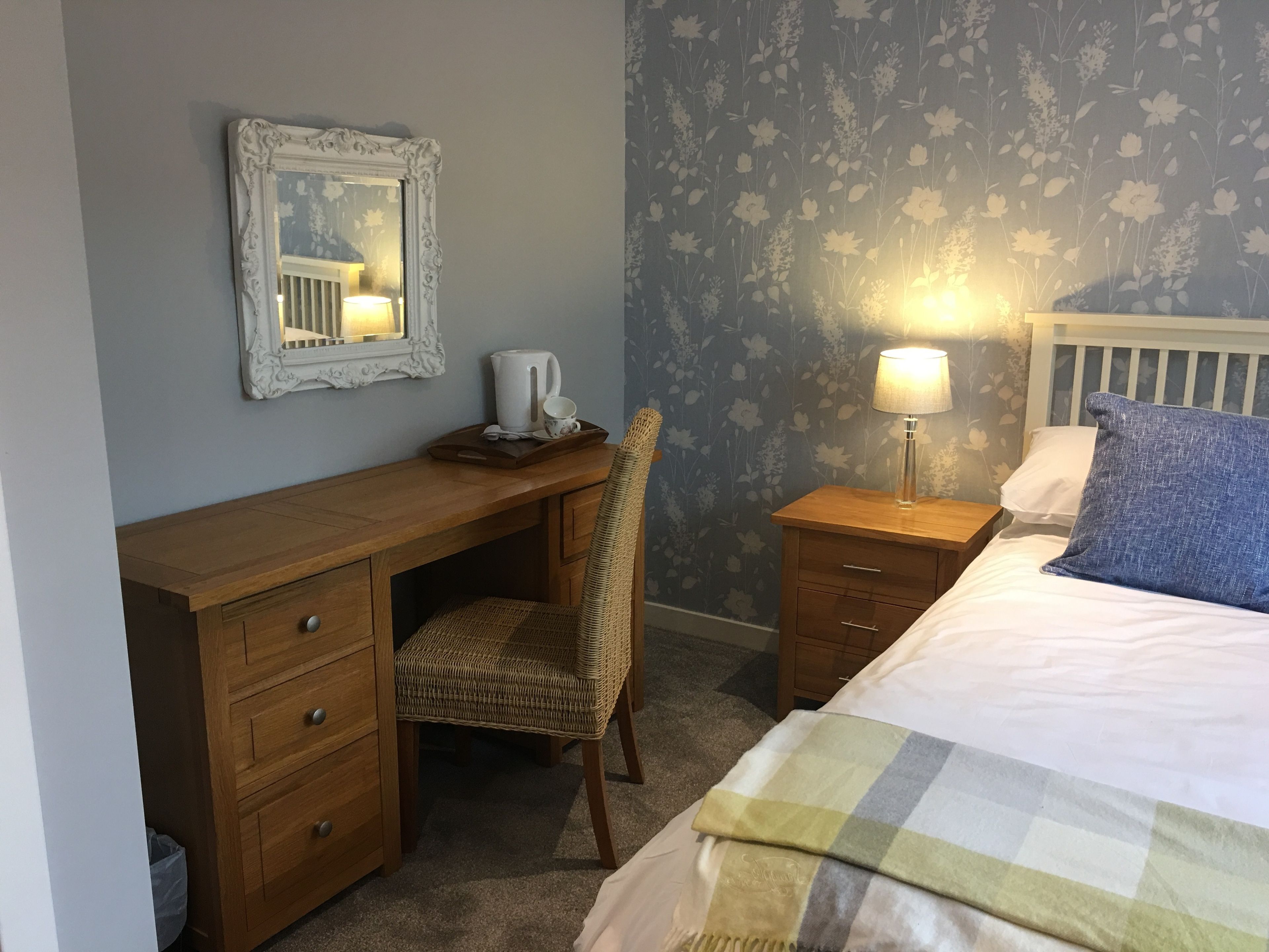 A bedroom with a wooden desk, a chair, a bedside table with a lamp, a bed with a blue pillow, and a floral wallpaper.