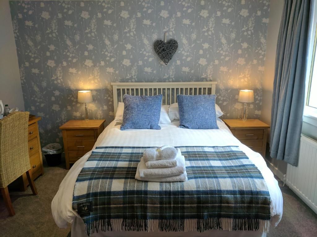 A hotel room with a bed, two bedside tables, a lamp, a heart-shaped decoration on the wall, and a window with curtains.