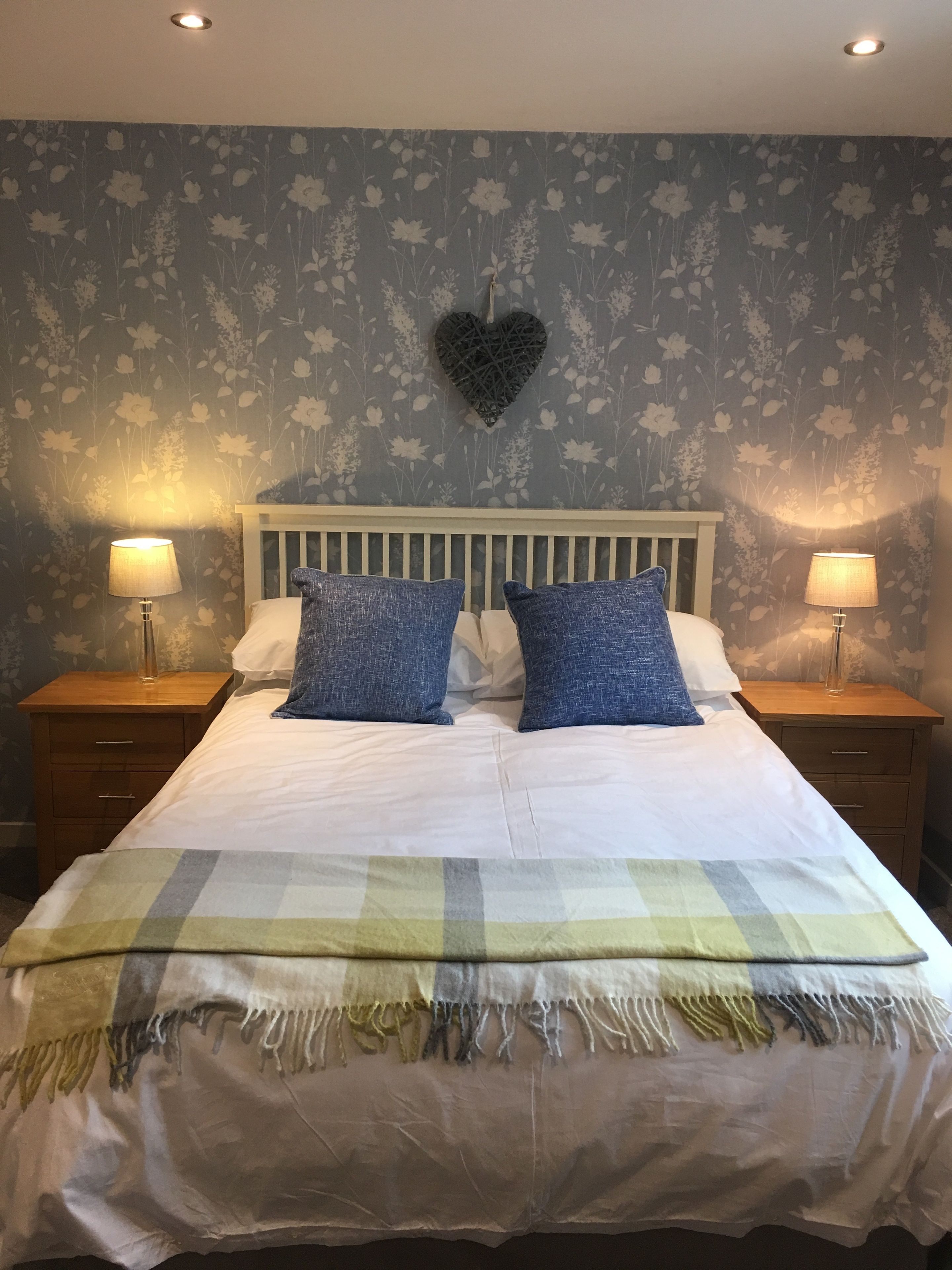 A bedroom with a bed, two nightstands, a heart-shaped wall decoration, and floral wallpaper.