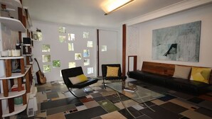 Una sala de estar moderna con un sofá negro, una mesa de centro de vidrio y un sillón negro. Hay varias ventanas con divisiones cuadradas, una guitarra y una estantería.