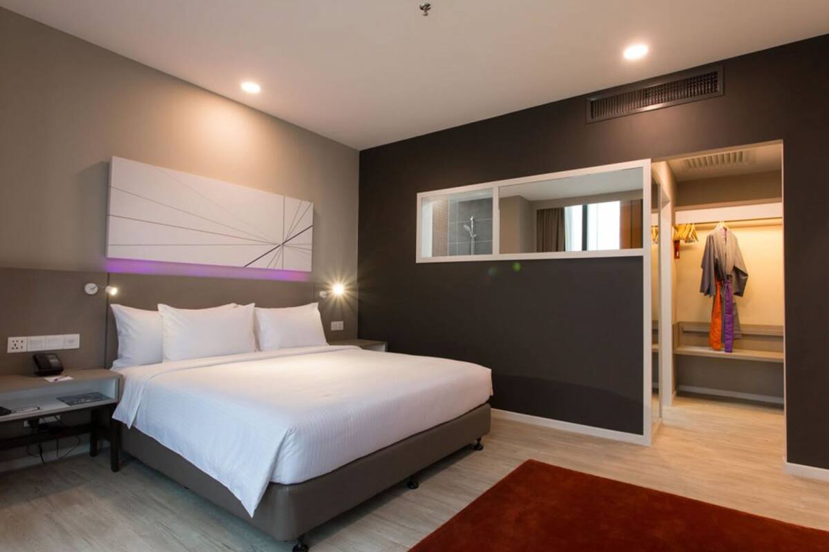 A modern bedroom with a large bed, a bedside table, a nightstand, a wall-mounted light, a mirror, and a closet with hanging clothes.