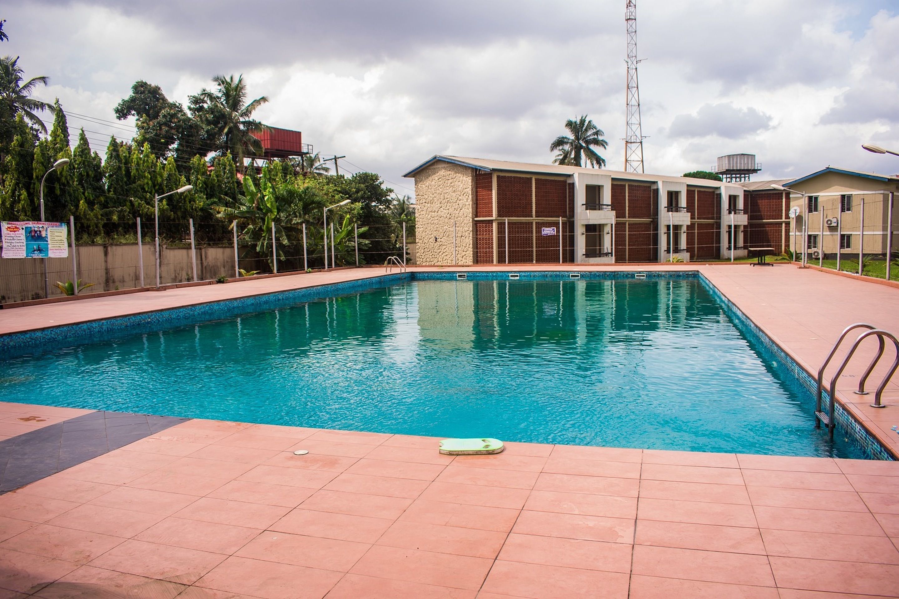 Une piscine rectangulaire, bordée d’une terrasse carrelée, entourée de bâtiments et de palmiers.