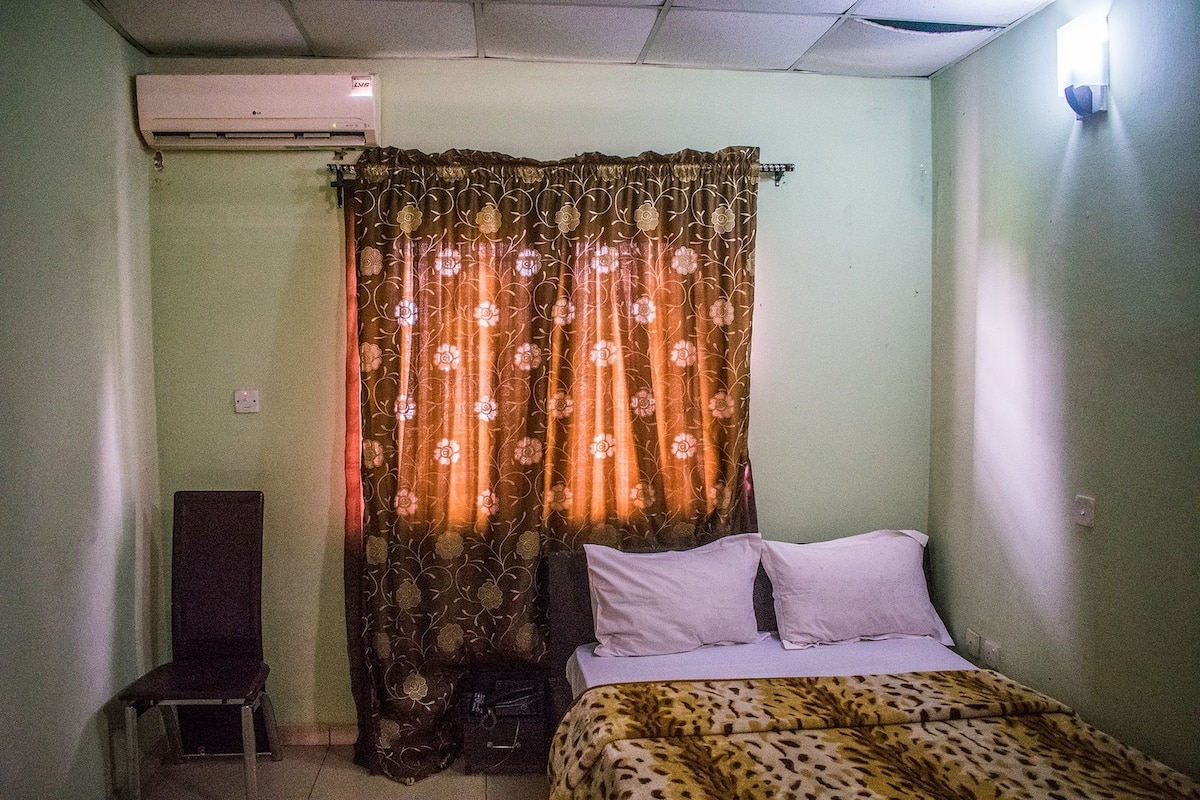 A room with a bed, a chair, a curtain, and an air conditioning unit.
