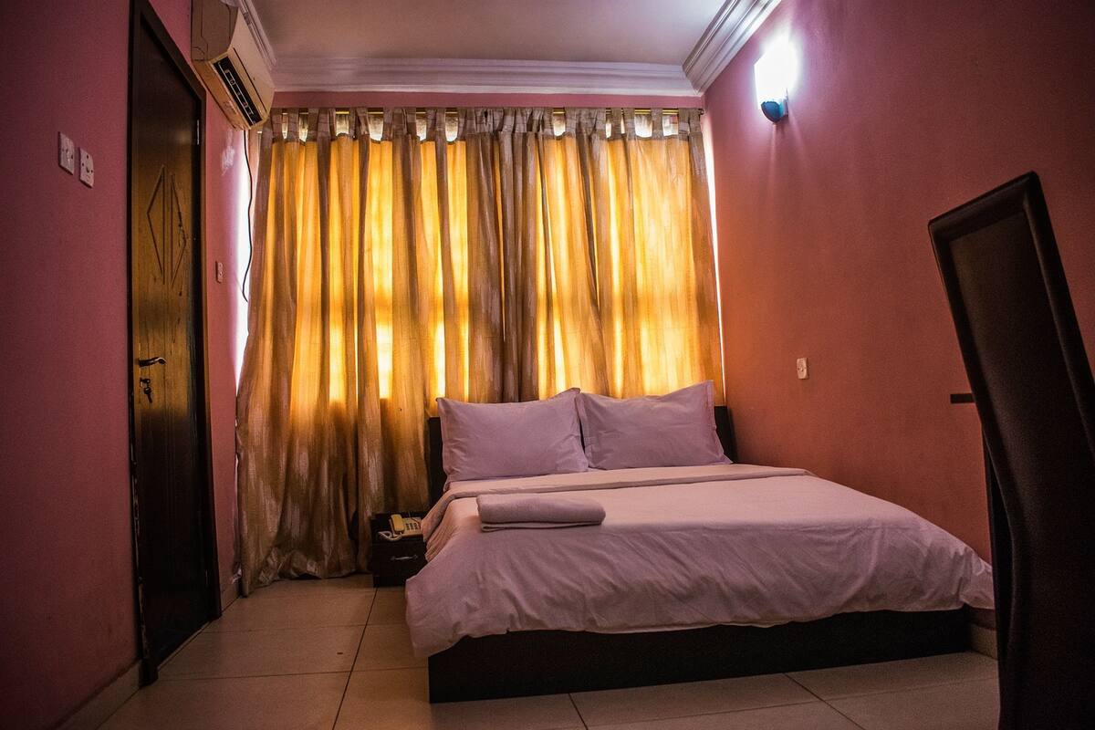 A bedroom with a bed, curtains, a nightstand, and a wall-mounted light.