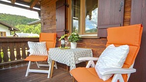 Une terrasse en bois avec deux chaises blanches dotées de coussins orange, une petite table recouverte d’une nappe blanche et une plante en pot.