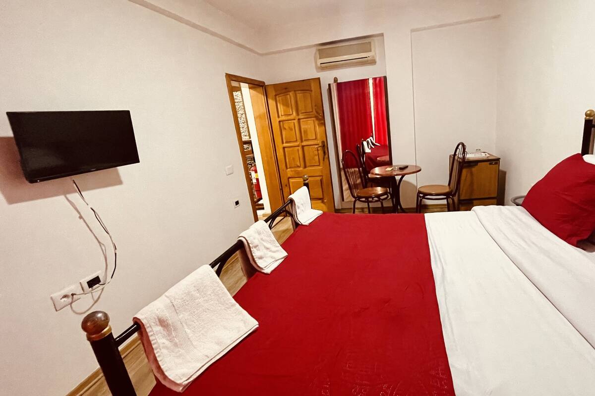 A hotel room with a bed, a TV mounted on the wall, a small table with chairs, and a door leading to a bathroom.