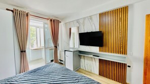 Un dormitorio con cama, televisor montado en una pared de paneles de madera, un escritorio pequeño y una ventana con cortinas.