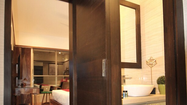 A hotel room with a wooden floor, a bed, a desk, a chair, a large mirror, and a bathroom with a sink and a potted plant. - Theory9 Premium Serviced Apartments Khar (Mumbai)
