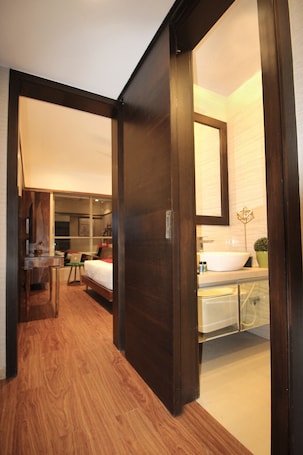 A hotel room with a wooden floor, a bed, a desk, a chair, a large mirror, and a bathroom with a sink and a potted plant.