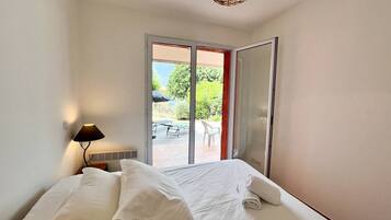 Ein Schlafzimmer mit Bett, Nachttisch, Lampe und einer Glastür, die auf eine Terrasse mit Tisch und Stühlen führt.