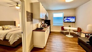 Ein kleines Hotelzimmer mit Bett, Küchenzeile, Essbereich und einem Fernseher, der ein Sportevent zeigt.