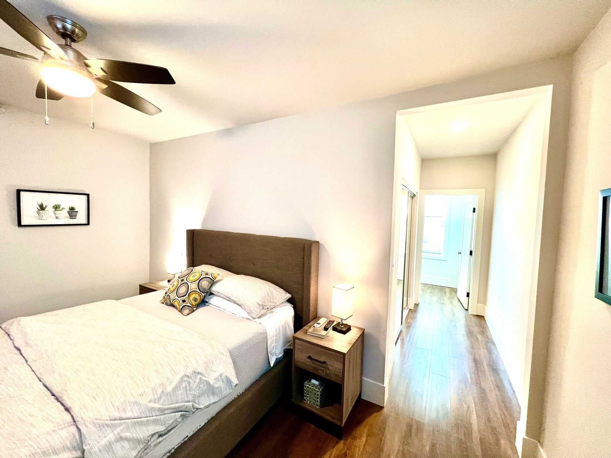A bedroom with a bed, nightstand, lamp, ceiling fan, and a framed picture on the wall.