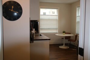 A compact kitchen with a round table and chairs, a clock, and a window with blinds.