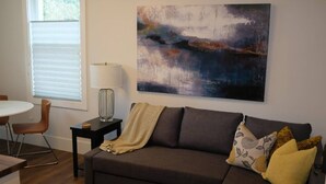 Un salon moderne avec un canapé marron foncé, un plaid à rayures et des coussins décoratifs. On y trouve une petite table d’appoint avec une lampe, et un tableau est accroché au-dessus.