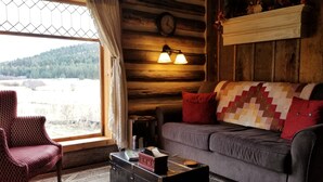 Interior acogedor de una cabaña con pared de troncos de madera, un sofá gris con colcha, una silla roja, una mesa con una maleta y una ventana con vistas a una zona boscosa.