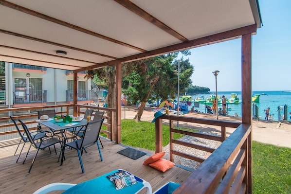 A covered outdoor seating area with a table and chairs, overlooking a beach with people and water activities. - Centinera Mobile Homes (Medulin)