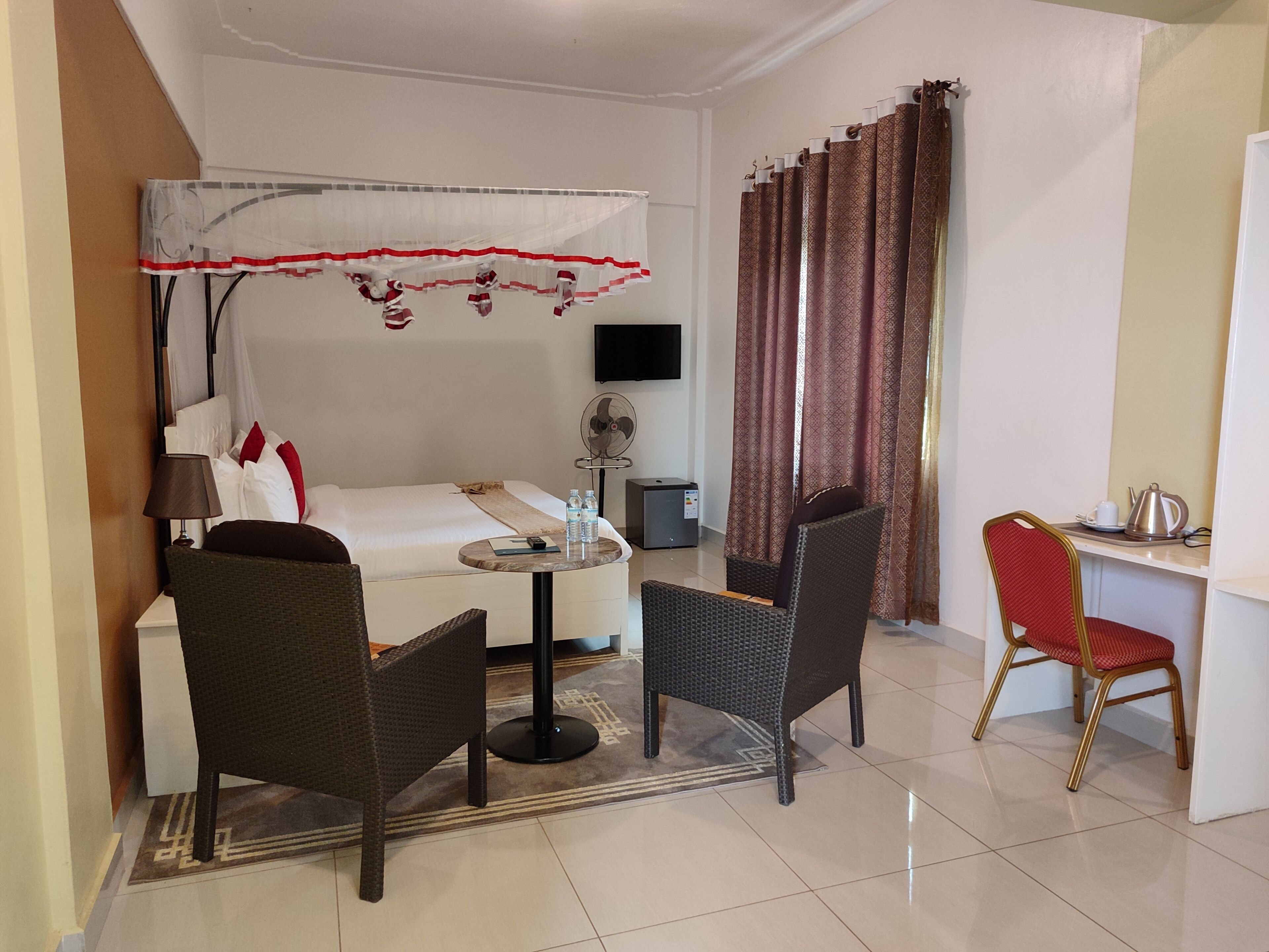 A hotel room with a bed, a desk, two chairs, a fan, a TV, and curtains.