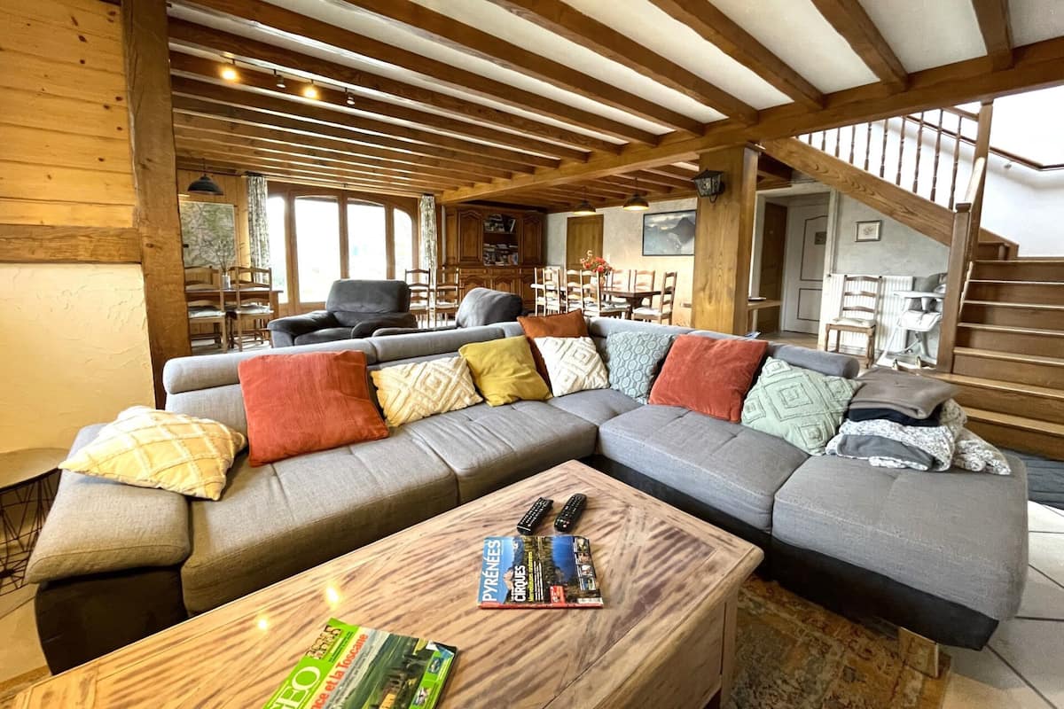 A cozy living room with a sectional sofa, wooden beams, a dining area, and a staircase.