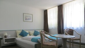 Quarto de hotel com cama, duas cadeiras, uma mesinha, mesa de cabeceira e janela com cortinas.