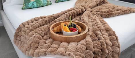 A neatly made bed with a brown blanket, a bowl of fruit and croissants, and decorative pillows with a green leafy pattern.