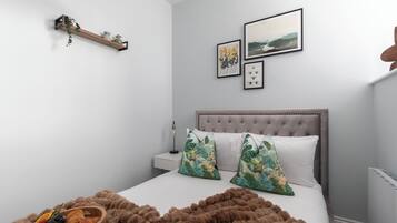 A bedroom with a tufted headboard, a bedside table with a lamp, a wall-mounted shelf with a plant, and a framed picture above the bed.