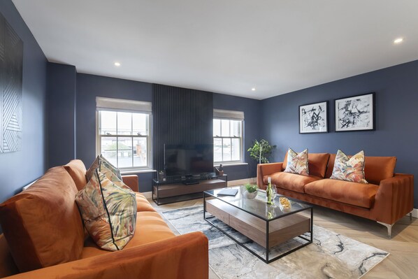 A modern living room with a large flat-screen TV, two sofas with orange cushions, a glass coffee table, and artwork on the walls. - Elliot Oliver - Luxury 3 Bedroom Town Centre Apartment With Parking (Cheltenham)