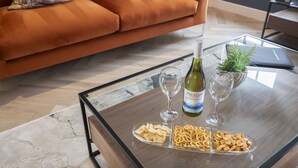 A modern living room with a glass coffee table, a bottle of wine, two wine glasses, and a plate of snacks. - Elliot Oliver - Luxury 3 Bedroom Town Centre Apartment With Parking (Cheltenham)