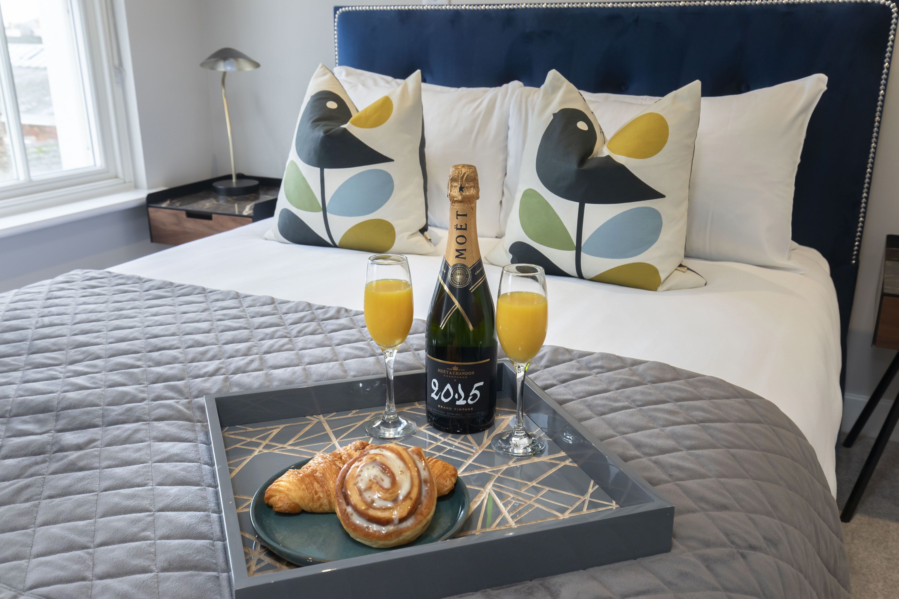 Una cama con cabecera azul, almohadas blancas con diseños de pájaros, una bandeja con una botella de champán, dos vasos de zumo de naranja y un croissant.