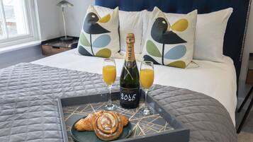 En säng med en blå sänggavel, vita kuddar med fågelmotiv, ett brickbord med en flaska champagne, två glas apelsinjuice och en croissant.