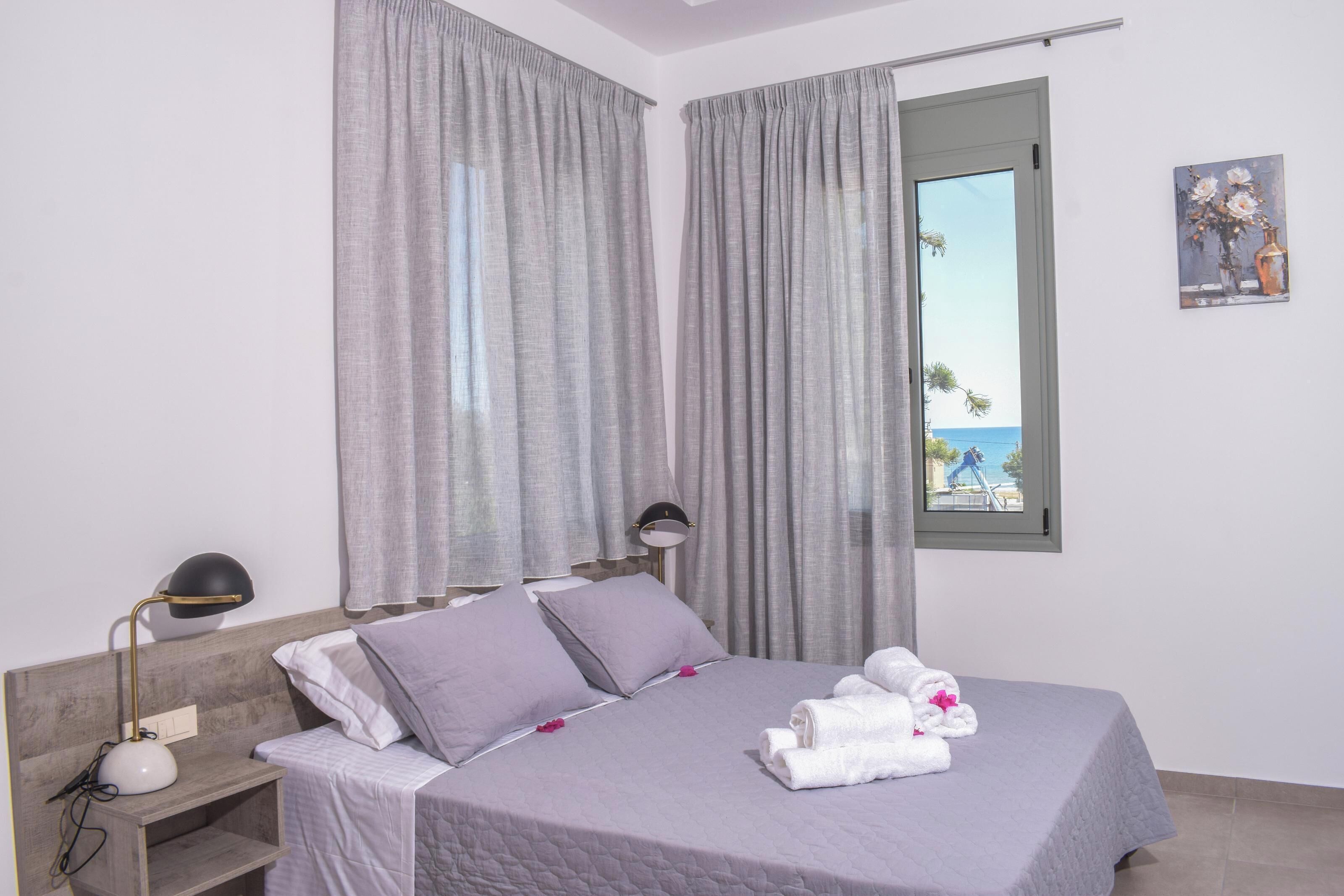 Una cama bien hecha con almohadas, una lámpara de noche y vista al mar a través de una ventana.