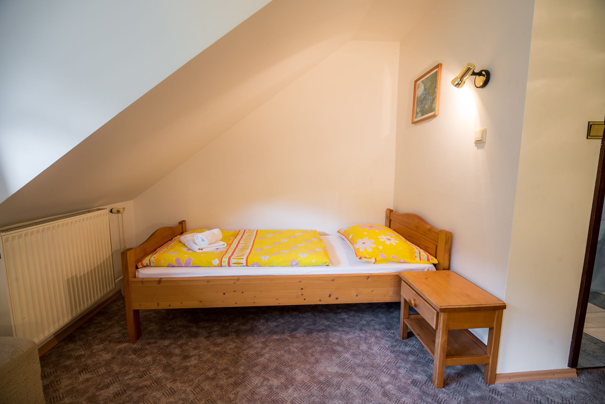 A small, single-bed room with a sloped ceiling, a wooden bed frame, a bedside table, and a radiator.