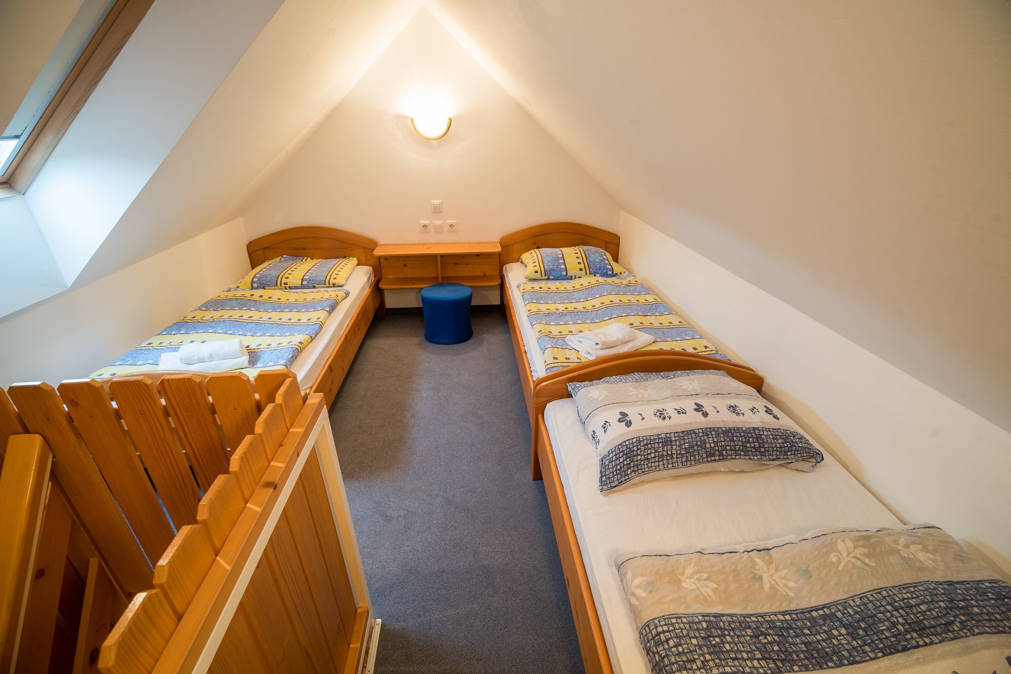 Une petite chambre mansardée de forme triangulaire, avec deux lits simples, un banc en bois et une petite table.