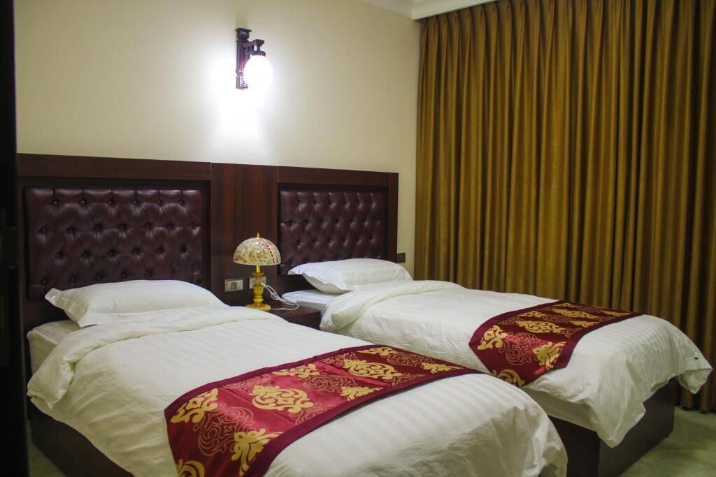 A hotel room with two beds, a nightstand, a lamp, and a large window with curtains.