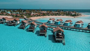 Cabañas sobre el agua conectadas por un muelle, con agua turquesa cristalina y una playa al fondo.