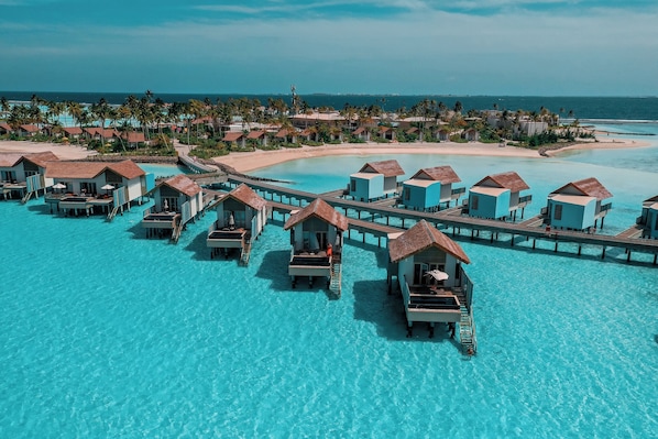 Overwater bungalows connected by a pier, with clear turquoise water and a beach in the background. - Hard Rock Hotel Maldives - FREE shared speedboat for stays from 01st Mar to 21st Dec 2026 (Emboodhoo Lagoon)
