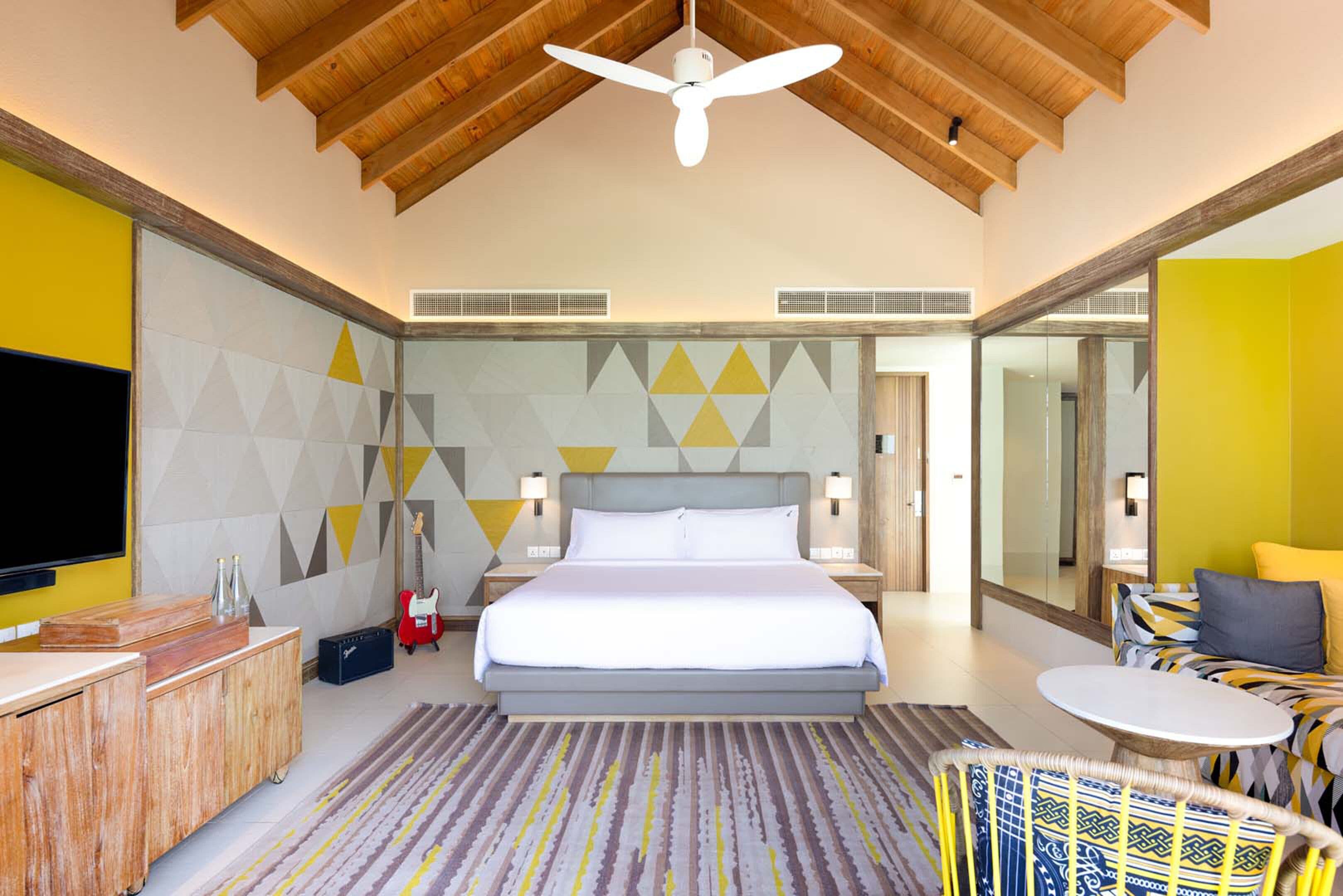 A modern hotel room with a large bed, a ceiling fan, a TV, a small table, and a seating area with a patterned rug.