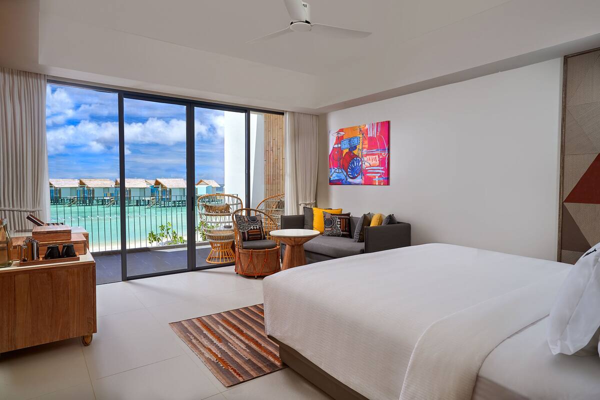 Une chambre d’hôtel avec un grand lit, une table à manger, une chaise, une petite table et une grande fenêtre donnant sur la plage.
