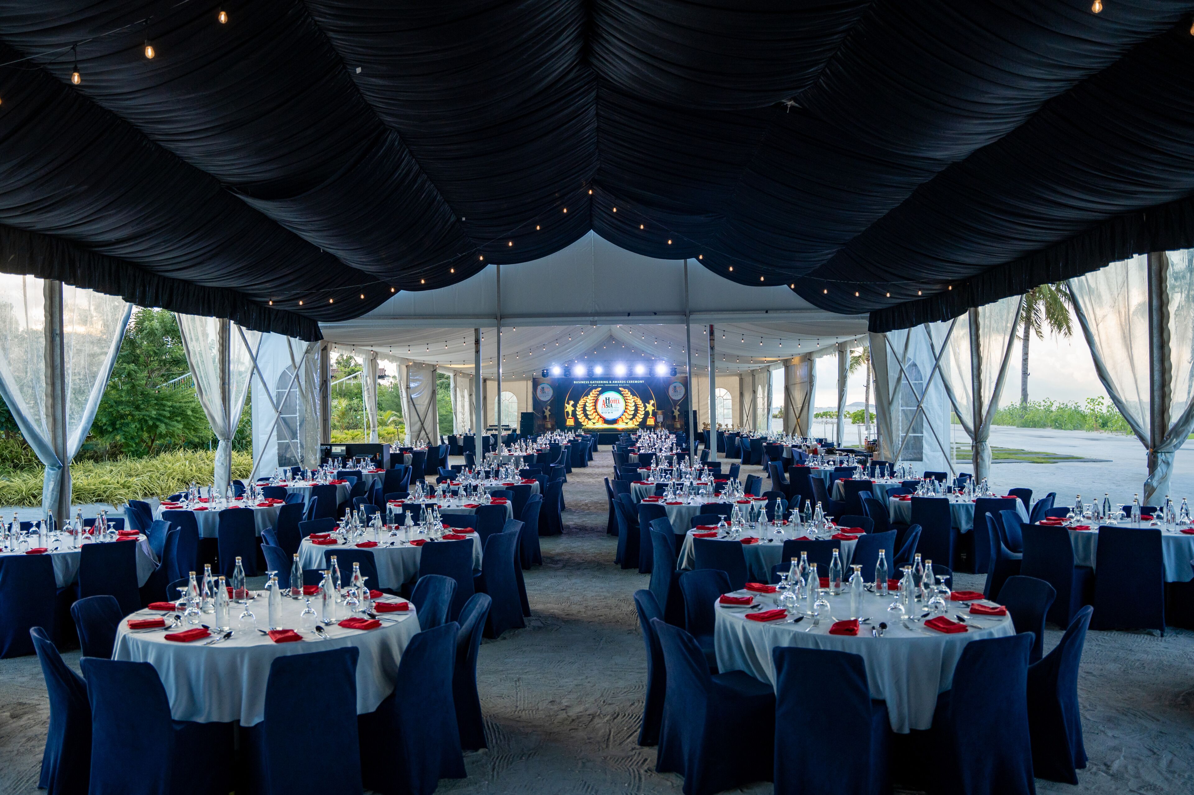 A tented dining area with round tables set for a formal event, featuring a central stage with lighting.