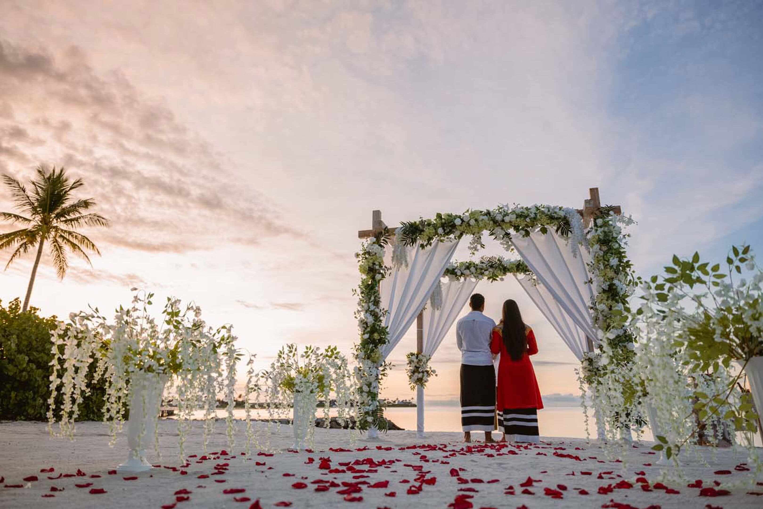 A beach wedding ceremony with a white arch decorated with flowers, palm trees, and a sunset in the background.