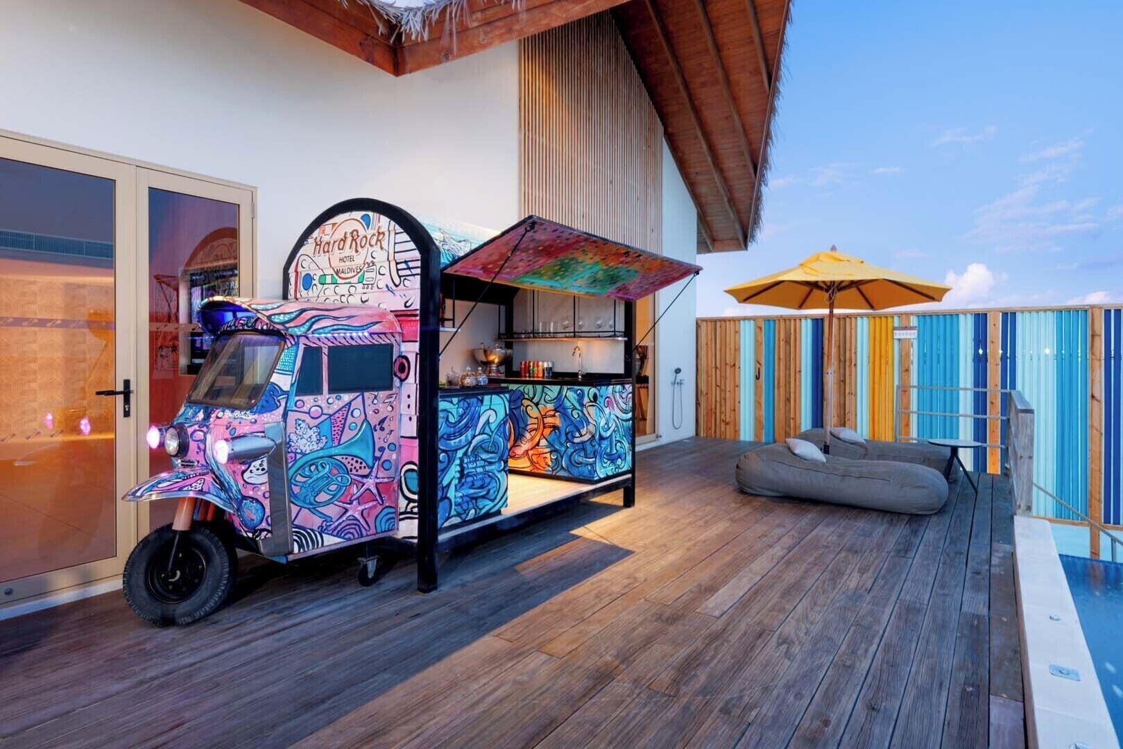 Un colorato furgone ristorante con un cartello che recita 'Hard Rock Hotel Maldives' parcheggiato accanto a un pontile di legno con piscina e una recinzione colorata.