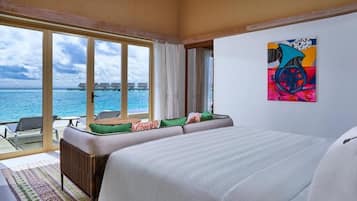 Habitación de hotel con una cama grande, vista al océano, un rincón de comedor con sillas y una obra de arte colorida en la pared.
