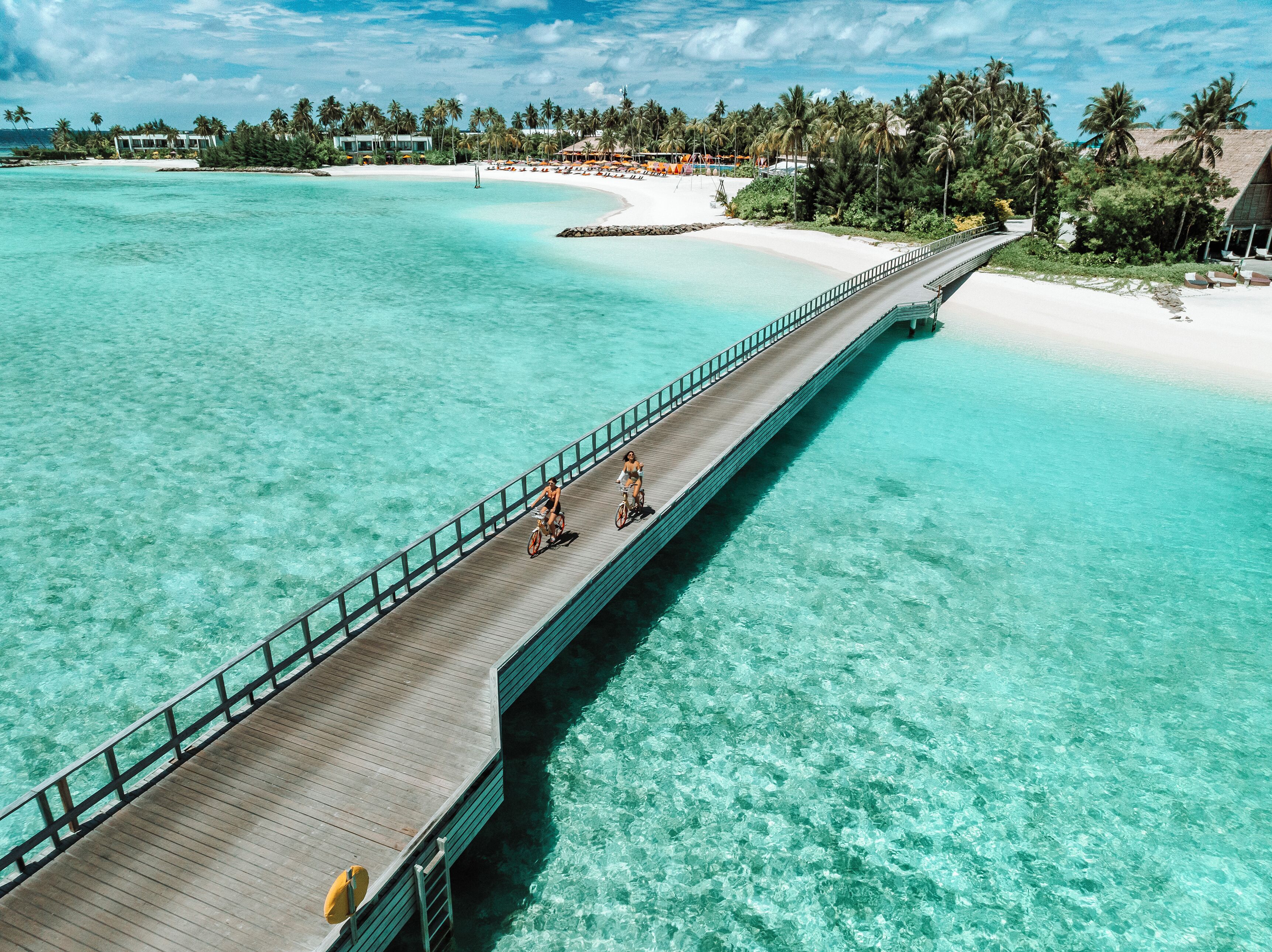 Een houten steiger die zich uitstrekt over turquoise water, naar een strand met palmbomen en resortgebouwen.