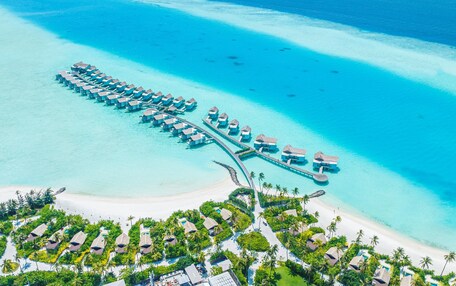 Vista aérea de un resort con bungalows sobre el agua dispuestos en forma de media luna, conectados por un puente, y rodeados de aguas turquesas cristalinas y una playa de arena.