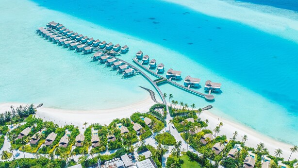 Vista aérea de un resort con bungalows sobre el agua dispuestos en forma de media luna, conectados por un puente, y rodeados de aguas turquesas cristalinas y una playa de arena.
