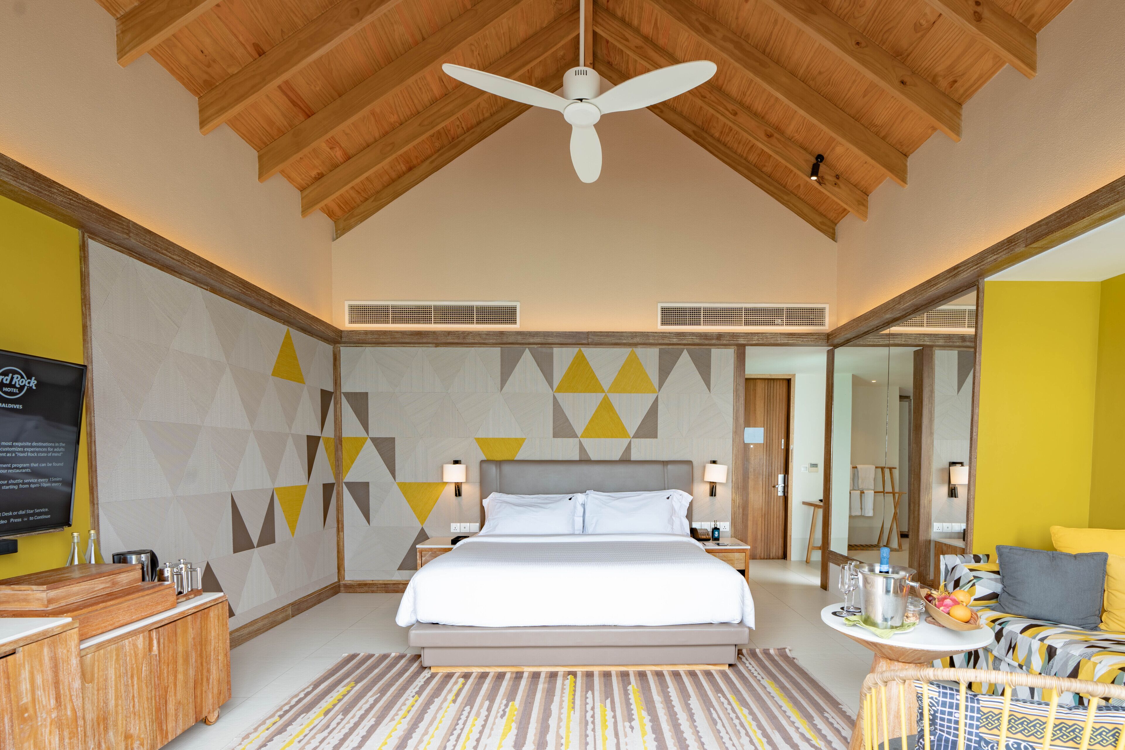 A modern hotel room with a large bed, wooden ceiling, geometric wall design, and a small side table with amenities.