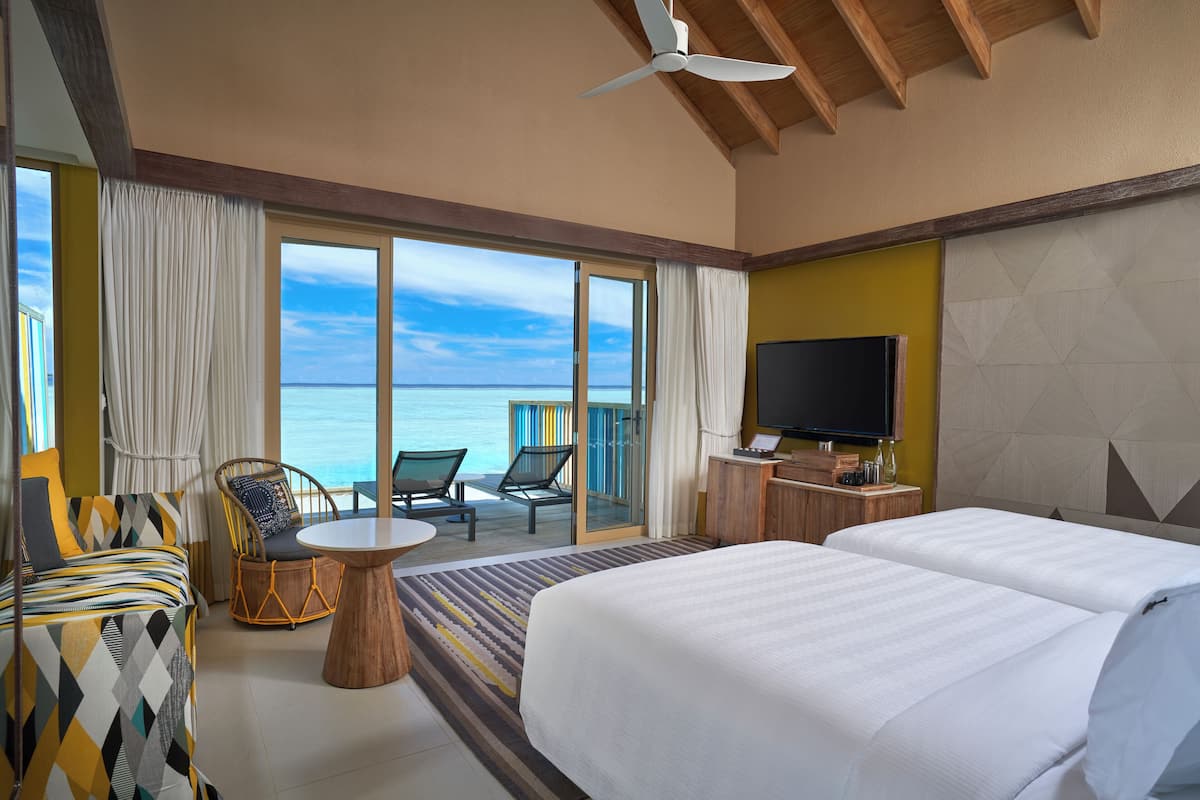 Une chambre d’hôtel dotée d’un grand lit, d’une télévision, d’un balcon avec vue sur la mer et d’un coin repas avec des chaises et une table.