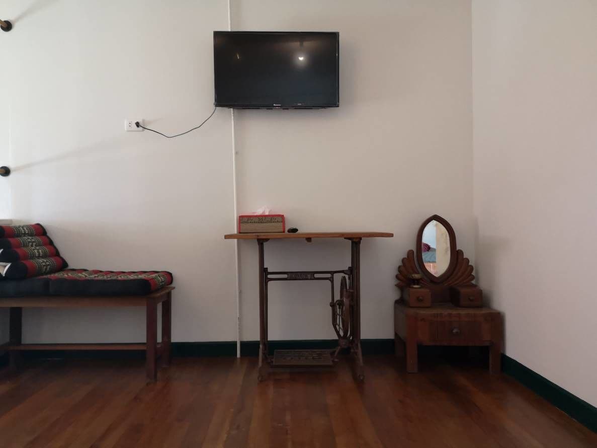 A room with a wooden floor, a sofa with patterned cushions, a sewing machine on a table, a television mounted on the wall, and a small wooden cabinet with a mirror.