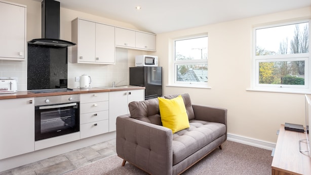 A compact kitchen with white cabinets, a black range hood, and a microwave. A grey sofa with a yellow pillow is in the living area, and there are two windows providing views of the outside. - Elliot Oliver - 2 Bedroom Town Centre Apartment (Cheltenham)